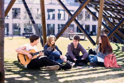 Elever på Pops Academy sitter tillsammans i gräset och lyssnar på när en av dem spelar gitarr.